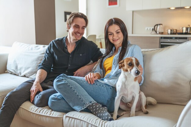 젊은 쾌활한 커플 남자와 여자는 부엌 배경에 아파트 집 소파에 앉아 개와 즐거운 시간을 보내고 있습니다.