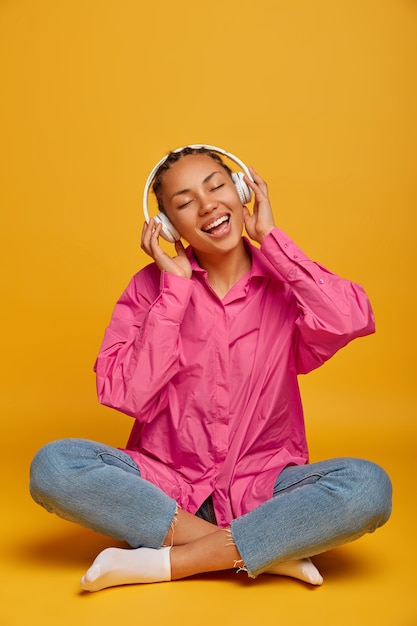 Free photo young cheerful ethnic woman enjoys music on floor, sits crossed legs, wears pink shirt, jeans and socks, listens audio track with loud sound, isolated on yellow wall, empty space above