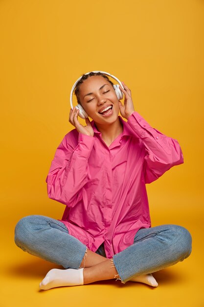 Молодая жизнерадостная этническая женщина наслаждается музыкой на полу, сидит скрестив ноги, носит розовую рубашку, джинсы и носки, слушает звуковую дорожку с громким звуком, изолирована на желтой стене, пустое место наверху