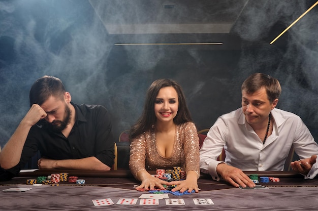 Молодые веселые друзья играют в покер в казино в дыму. Девушка выиграла, а парни проиграли. Молодежь делает ставки в ожидании крупного выигрыша. Азартные игры на деньги. Игры удачи.