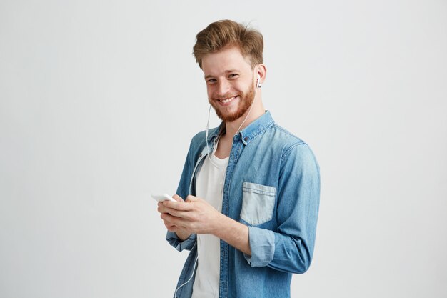 Young cheerful guy in headphones smiling holding phone listening to music.