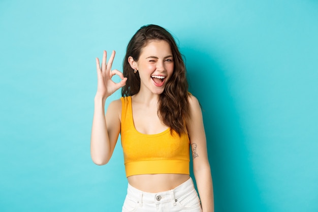 Young cheerful lady in summer clothes winking and saying yes, alright gesture, making OK sign in approval, standing against blue background.