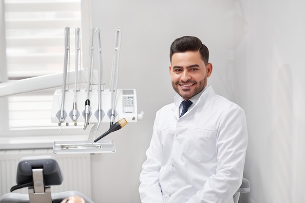 Young cheerful male professional dentist smiling joyfully sitting at his office