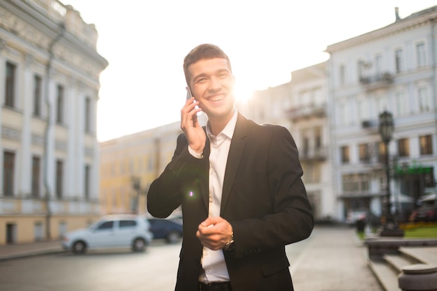 ファジーな背景に美しい街の景色と携帯電話で楽しく話している黒いジャケットと白いシャツの若い陽気な男