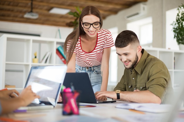 Молодой веселый мужчина в рубашке и женщина в полосатой футболке и очках работают вместе с ноутбуком Креативные деловые люди проводят время на работе в современном уютном офисе