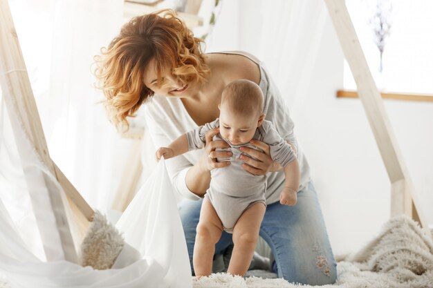 Young cheerful mother laughing and playing with newborn cute son in cozy light bedroom in morning. Atmosphere of happy family life.