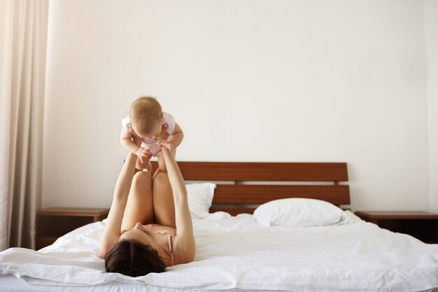 Young cheerful mother playing with her newborn baby lying on bed at home.
