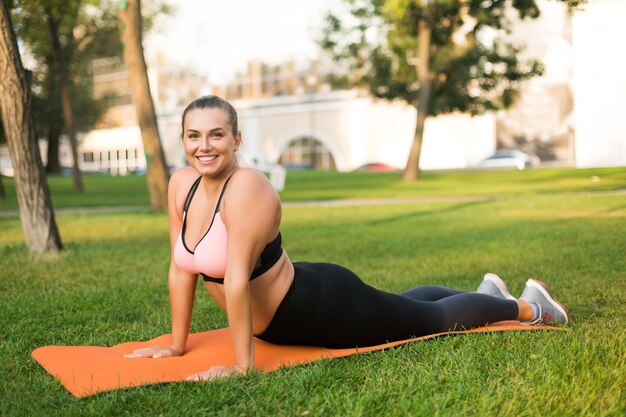 Молодая веселая женщина больших размеров в спортивном топе и леггинсах, практикующая йогу на оранжевом коврике для йоги, радостно смотрит в камеру, проводя время на зеленой траве в парке