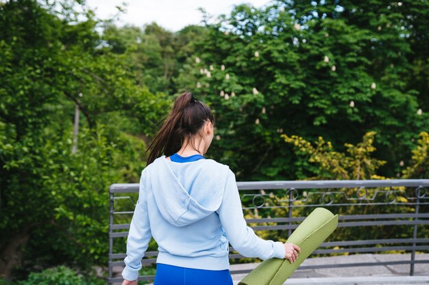 Молодая веселая спортивная женщина, идущая в городском парке, держащем фитнес-коврик.