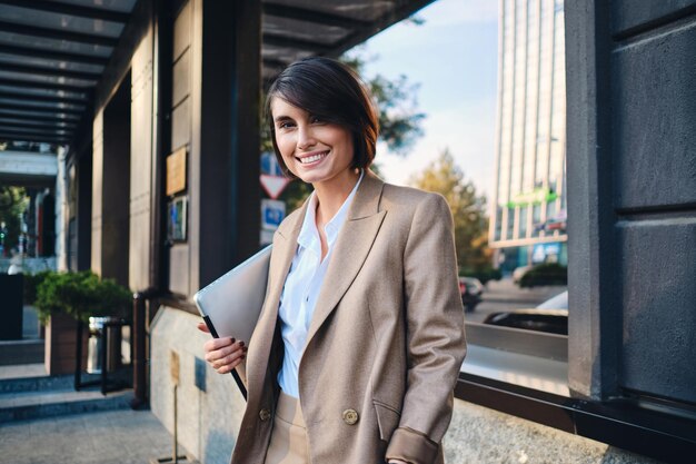 Young cheerful stylish businesswoman with laptop happily looking in camera on city street