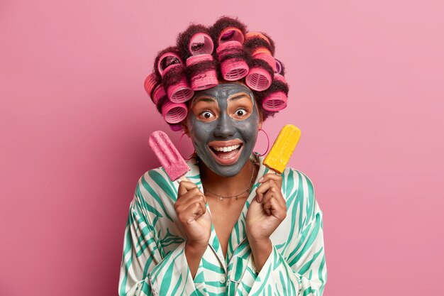 Young cheerful woman applies clay mask for healthy skin and reducing fine lines, poses with delicious cold ice cream, does beauty peeling procedures for face, wears hair curlers, wears dressing gown