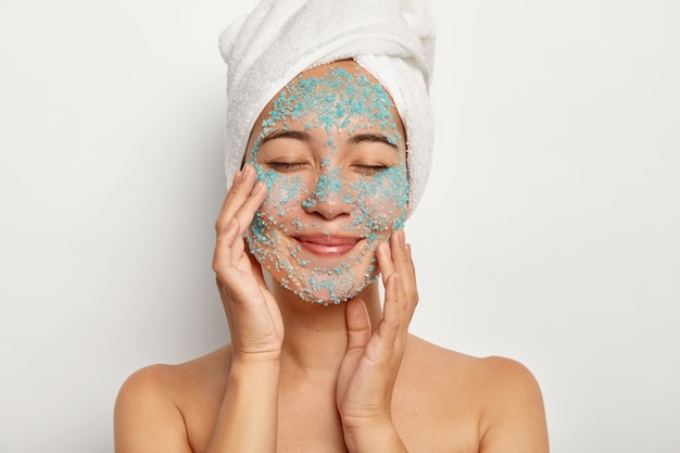 Young cheerful woman applies natural scrub on face, touches cheeks, keeps eyes closed, wears towel, has beauty procedures after taking shower, models indoor. Female model with blue sea salt on skin