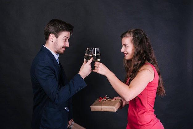 Young cheerful woman clinging glasses of drink with man and holding present boxes