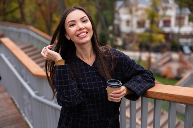 Young cheerful woman in coat with coffee to go happily looking in camera in park