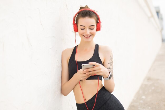 짙은 회색 스포티 상의와 빨간 헤드폰을 쓴 레깅스를 입은 젊고 쾌활한 여성은 야외에서 음악을 들으면서 휴대폰으로 행복하게 벽에 기대어 있습니다