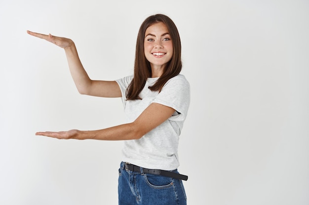 Молодая веселая женщина демонстрирует большой знак или баннер, формируя продукт большого размера на белой стене и улыбаясь