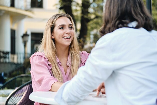 ピンクのドレスを着た若い陽気な女性は、屋外のカフェでロマンチックなデートで彼氏を喜んで見ています