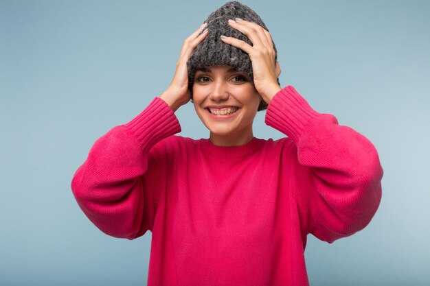 ピンクのセーターと灰色のニット帽子の若い陽気な女性は、分離された青い背景の上のカメラで喜んで見ています