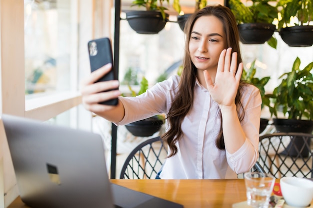 友達とチャットするためにスマートフォンで自分自身を撮影しながらポーズをとる若い陽気な女性、カフェに座っている間携帯電話で自画像を作る魅力的な笑顔の流行に敏感な女の子