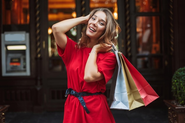 Young cheerful woman in red dress joyfully looking in camera while holding colorful shopping bags in hand with big door on background