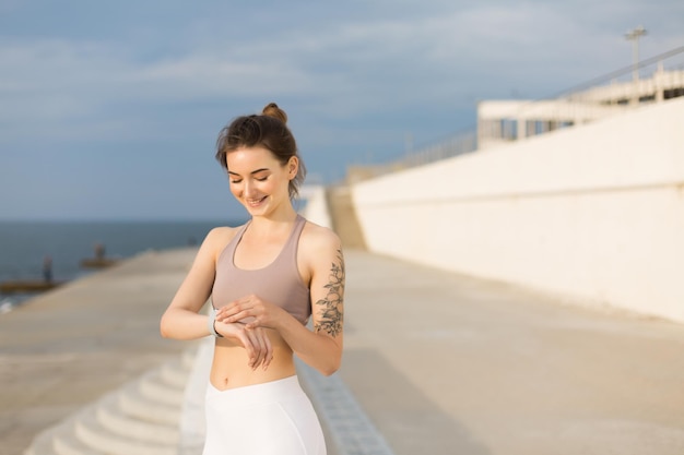 Young cheerful woman in sporty top happily looking on hand watch while spening time outdoor near the sea