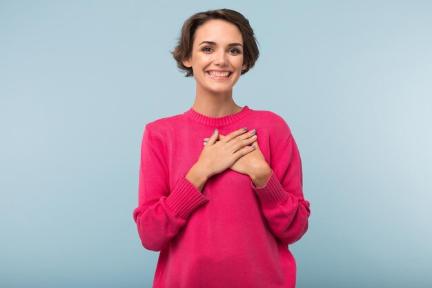 Young cheerful woman with dark short hair in pink sweater holding hands on chest while happily looking in camera over blue background