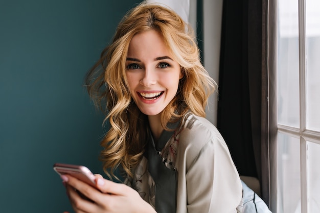 Young cheerful woman with long wavy hair with pink smartphone in hands looking and smiling, sitting next to window. Wearing silk pajama. Turquoise wall, curtains.