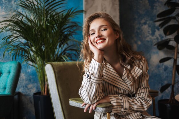 Молодая жизнерадостная женщина с волнистыми волосами в полосатом плаще, опираясь на руку, счастливо смотрит в камеру, проводя время в современном кафе