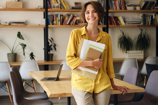 Free photo young cheerful woman in yellow shirt leaning on desk with notepad and papers in hand while joyfully looking in camera in modern office