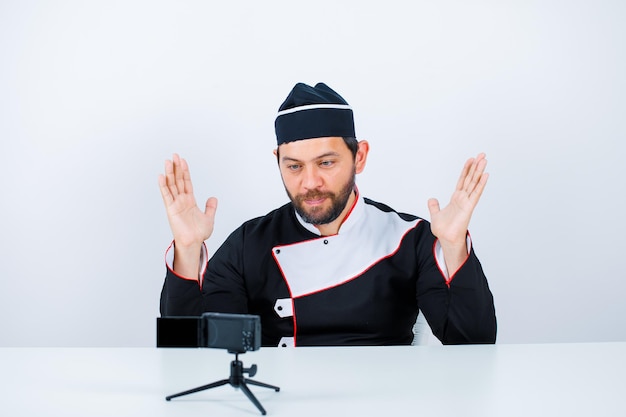 Young chef blogger is posing to his mini camera by raising up his hands on white background