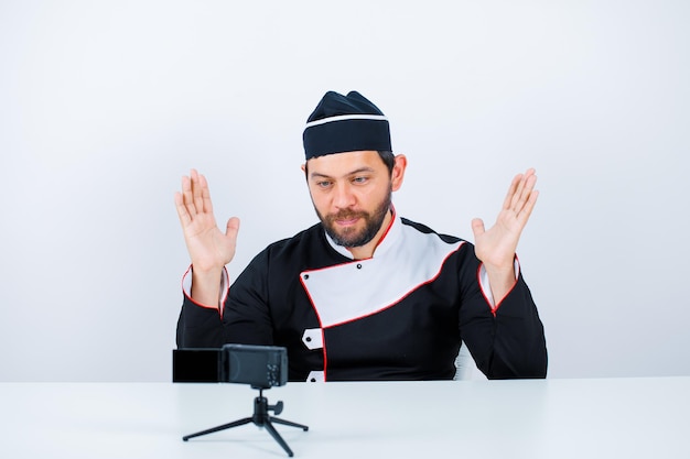 Free photo young chef blogger is posing to his mini camera by raising up his hands on white background