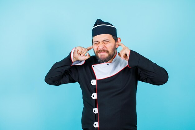 Young chef is closing eyes an dcovering eras with forefingers on blue background