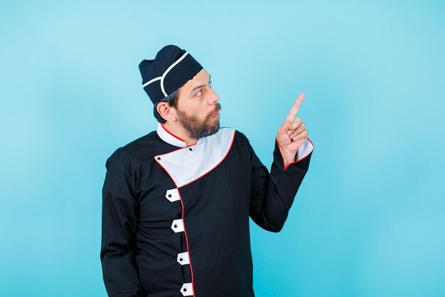 Young chef is looking up and pointing up with forefinger on blue background