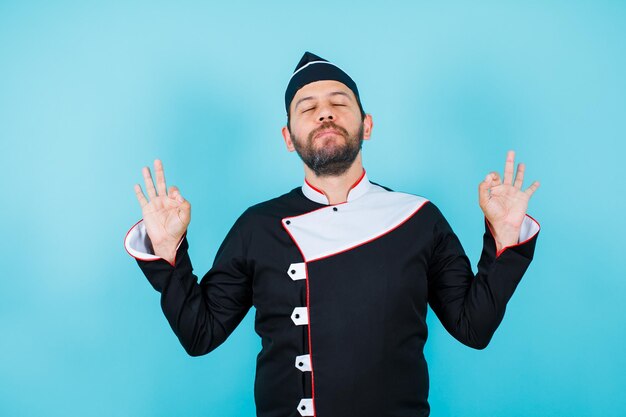 Young chef is meditating by closing eyes and raising up his hands on blue background