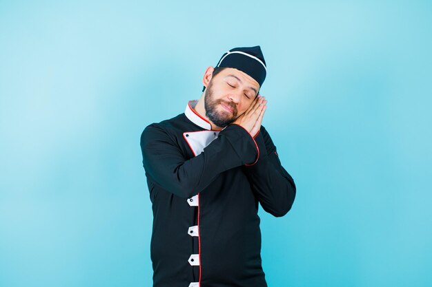 Young chef is showing sleeping gesture by holding hands together near cheek and closing eyes on blue background