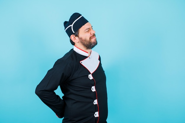 Young chef with back pain is putting hands behind back on blue background