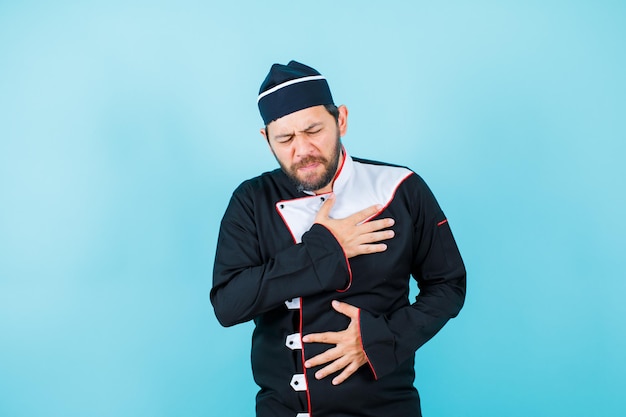 Young chef with pain is putting hand on chest on blue background