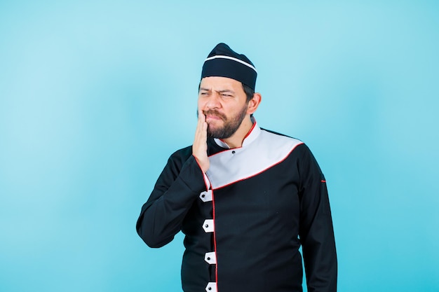 Young chef with toothache is putting hand on cheek on blue background