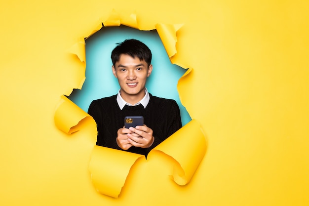 若い中国人男性は携帯電話を保持し、引き裂かれた黄色の壁の穴に頭を保ちます。破れた紙の男性の頭。