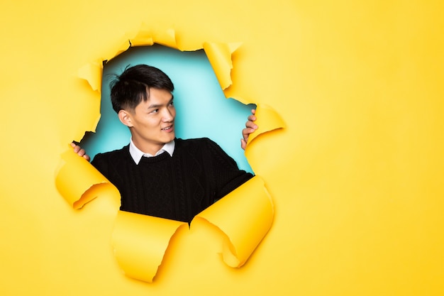 Young chinese man keeps head in hole of torn yellow wall. Male head in ripped paper.