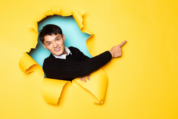 Young chinese man pointed side with hands keeps head in hole of torn yellow wall. Male head in ripped paper.