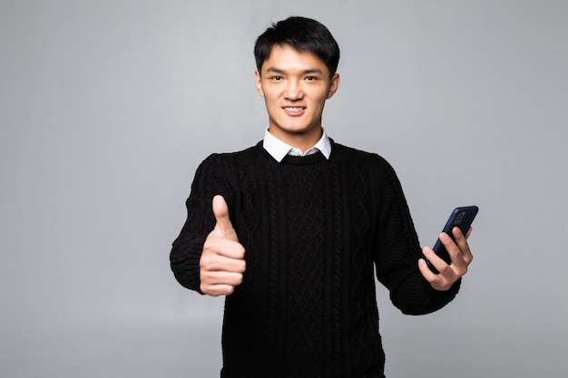 Young chinese man talking on the smartphone standing happy with big smile doing ok sign, thumb up with fingers, excellent sign over isolated white wall