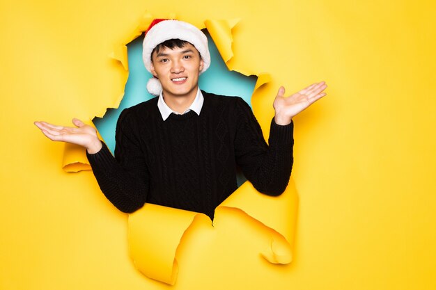Young chinese man with santa hat and raised hands keeps head in hole of torn yellow wall. Male head in ripped paper.