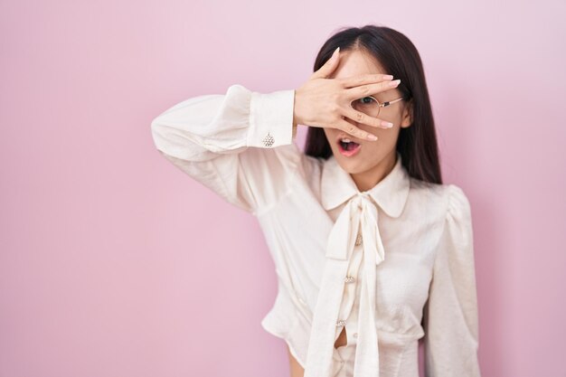 ピンクの背景の上に立っている若い中国人女性は、ショックで顔と目を手で覆って覗き込み、恥ずかしそうな表情で指を通して見ています。