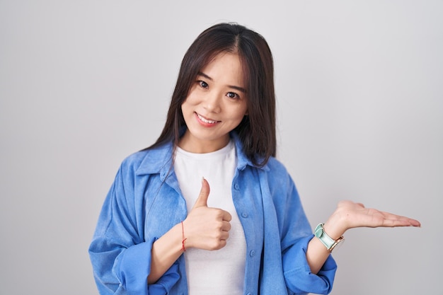 흰색 배경 위에 서서 손바닥을 보여주고 엄지손가락을 치켜들고 행복하고 쾌활한 미소를 짓는 젊은 중국 여성