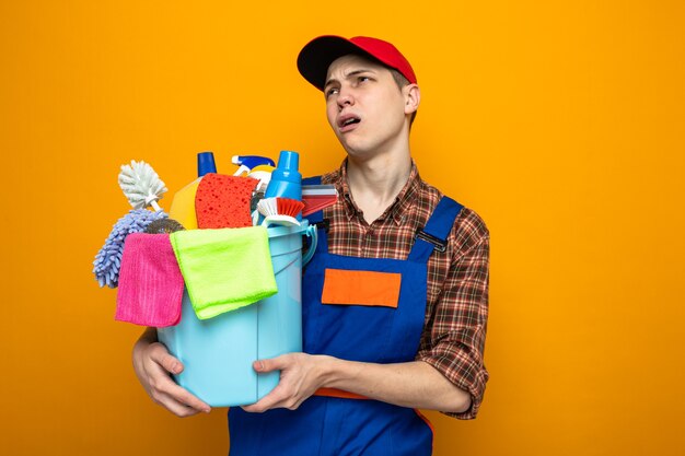 Молодой уборщик в униформе и кепке держит ведро с чистящими инструментами, изолированное на оранжевой стене