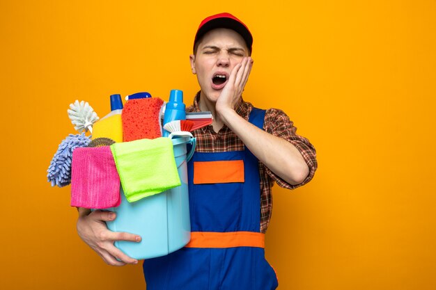 Young cleaning guy wearing uniform and cap holding bucket of cleaning tools isolated on orange wall