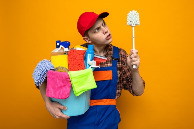 Young cleaning guy wearing uniform and cap holding bucket of cleaning tools and looking at brush in his hand isolated on orange wall