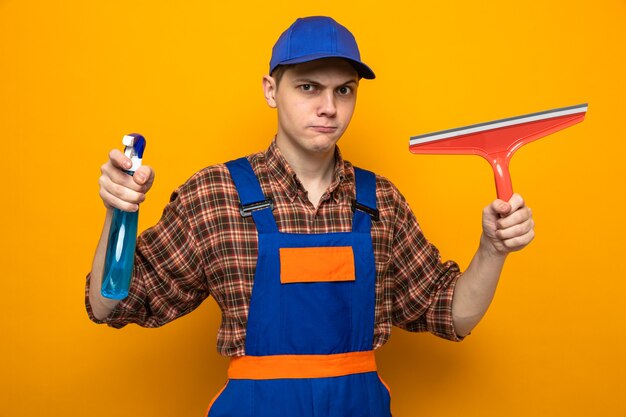 Young cleaning guy wearing uniform and cap holding cleaning agent with mop head isolated on orange wall