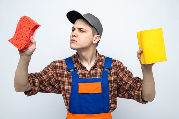 Young cleaning guy wearing uniform and cap holding and looking at sponges isolated on white wall